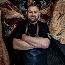 Keith Grant, master butcher, standing with his arms folded in a black leather apron in a dry-ageing room, sides of beef hanging on either side of him.