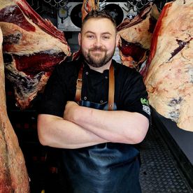 Master butcher Keith Grant, arms folded in his chill room with dry-aged beef carcasses hanging behind him, who leads the butcher's counter at Dublin Cookery School's steak classes.