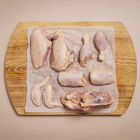 A whole chicken jointed into eight pieces on baking parchment on a wooden board — two breasts, two thighs, two drumsticks, two wings and the stripped carcass — photographed from above on a pale pink background.