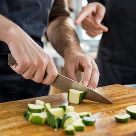 Student cutting courgette on a wooden board during a Knife Skills class at Dublin Cookery School, with a tutor's hands visible in the background guiding technique