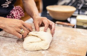 Hands shaping a loaf of bread dough on a floured board beside a proving basket.