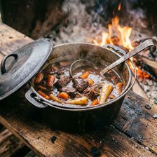 A well-used cast iron pot of beef and Guinness stew with slow-cooked beef, carrots and parsnips in a deep dark broth, photographed over an open fire with flames and rising steam, resting on a charred wooden surface outdoors.