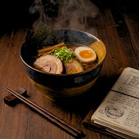A steaming bowl of ramen with chashu pork, soft-boiled egg, nori, and spring onions in a dark ceramic bowl, served on a wooden surface beside chopsticks and an open handwritten notebook.