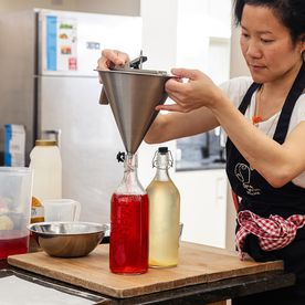 A Dublin Cookery tutor student in a black apron using a stainless steel funnel to bottle homemade red and yellow cordials on a wooden chopping board in a professional kitchen.