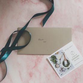 A Dublin Cookery School gift voucher in a gold envelope addressed to Mum, presented on a pink marble surface with a teal satin ribbon and a branded DCS card.