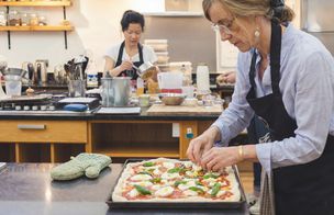 A woman in a blue shirt and glasses adds fresh mozzarella and basil to a rectangular pizza dough in the professional kitchen of Dublin Cookery School. In the background, another student in a black apron stirs a saucepan on a counter equipped with mixers and kitchen utensils. The scene demonstrates the hands-on learning and technique-led environment of a Gourmet Pizza Masterclass.