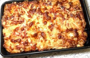 A woman in a blue shirt and glasses adds fresh mozzarella and basil to a rectangular pizza dough in the professional kitchen of Dublin Cookery School. In the background, another student in a black apron stirs a saucepan on a counter equipped with mixers and kitchen utensils. The scene demonstrates the hands-on learning and technique-led environment of a Gourmet Pizza Masterclass.