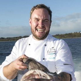 Fishmonger Scott Smullen laughing as he holds a large monkfish by the shore, in a BIM Young Fishmonger chef's jacket — guest fishmonger at Dublin Cookery School's seafood classes.