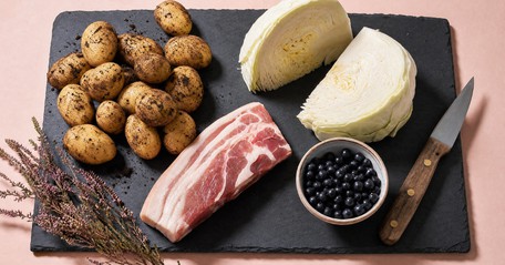 Raw ingredients for the traditional Irish Lúnasa meal arranged on a dark slate board — freshly dug new potatoes with soil still on their skins, a piece of bacon collar, a quartered white cabbage and a small bowl of dark fraughans, with a sprig of heather.