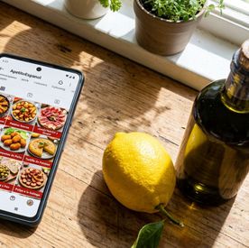 A phone on a sunlit windowsill showing an Instagram grid of Spanish dishes, beside a lemon and a bottle of olive oil.
