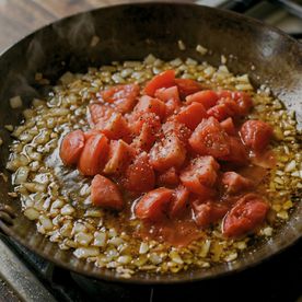 Chopped tomatoes added to softened onion and garlic in a dark pan on the hob — the start of a sofrito base, a foundational cooking technique taught at Dublin Cookery School.