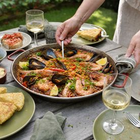 A seafood paella with prawns and mussels being served on a garden table, with tapas and white wine.