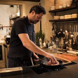 Man in a dark apron pressing a steak into a hot cast iron pan, steam rising, during the Steaks, Sauces and Sides class at Dublin Cookery School