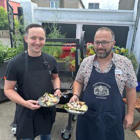 A man in a striped butcher's apron and glasses working at a heavy iron grill outdoors, pressing down on burger patties with a spatula while a woman in a navy striped jumper watches closely.