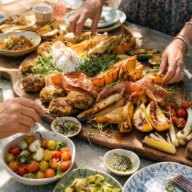 A large wooden board with a summer feast spread including crab cakes, blistered melon and pear with prosciutto and charred vegetables, hands reaching in to serve — Summer BBQ Feast at Dublin Cookery School