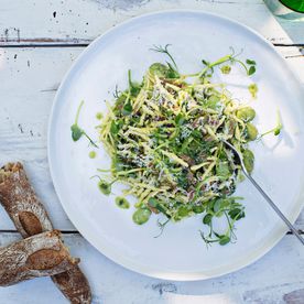 A plate of trofie pasta with broad beans, pea shoots, fresh herbs and shaved hard cheese, served with crusty bread on a white wooden table.