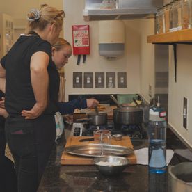 A female instructor supervising a young student cooking at a gas hob during a teen cooking class, with a fire blanket mounted on the wall in the background