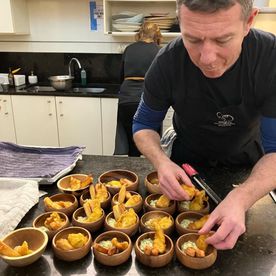 A Dublin Cookery School tutor plating fried pastry bites into small wooden serving bowls in the Blackrock kitchen, rows of completed portions lined up on the worktop.